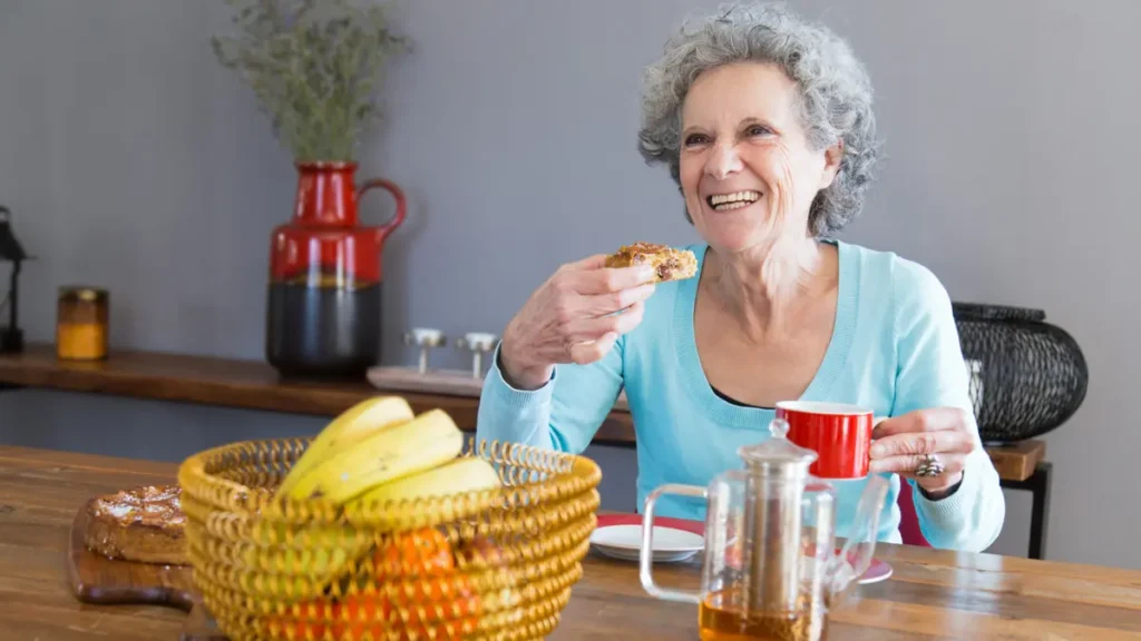 Happy senior woman laughing at kitchen table enjoying a meal, representing the natural appetite satisfaction from resistant starch foods.