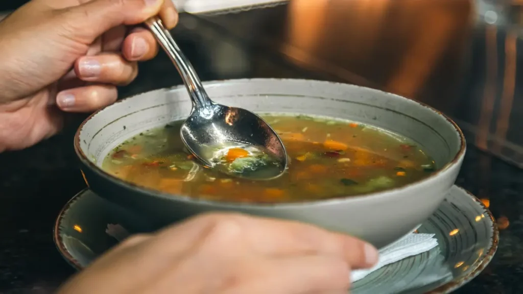 A person holding a silver spoon in a bowl of warm vegetable broth made from dissolved bouillon to create a quick, savory meal.
