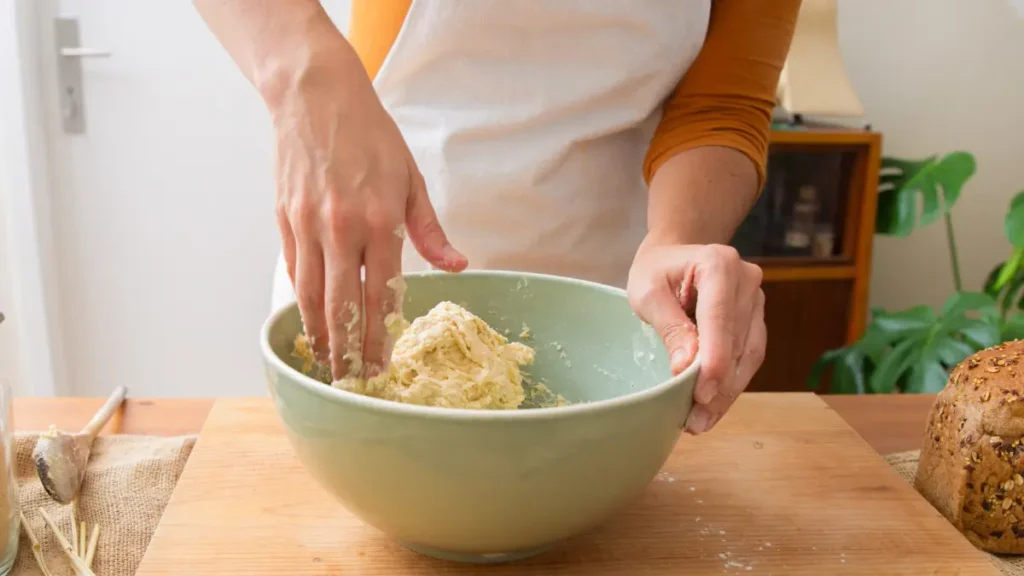 Mature hands mixing a shaggy and sticky no-knead bread dough in a green ceramic bowl.
