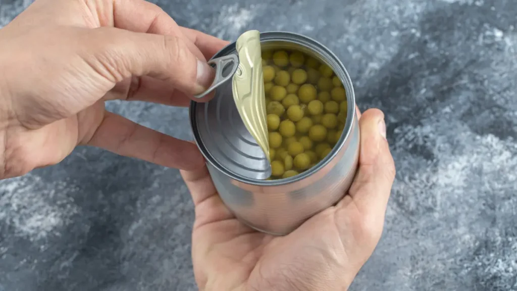 Hands pulling the metal tab to open a can of green peas preserved in liquid brine.
