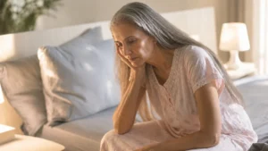 Exhausted woman over 50 sitting on bed edge in the morning, capturing the persistent fatigue and low energy caused by vitamin D deficiency.