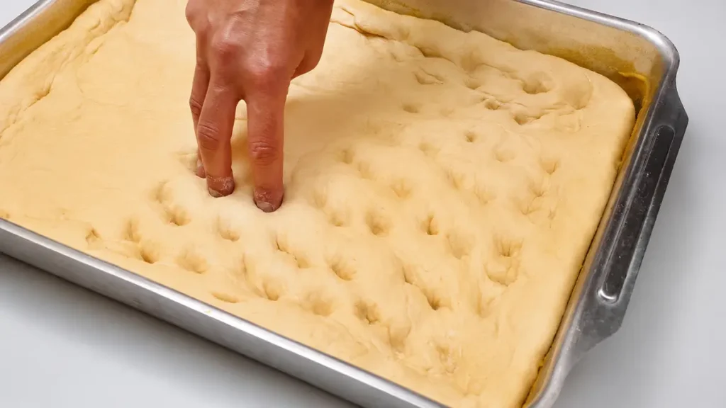 Hand pressing fingers into fully proofed bread dough on a baking sheet to test if it is ready to bake.
