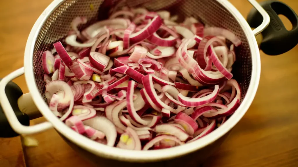 Sliced red onions resting in a metal colander ready to be cooked into a savory aromatic base.
