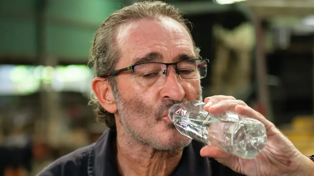 A mature man with glasses drinking from a water bottle in an industrial setting, symbolizing recovery from thirst and dehydration.