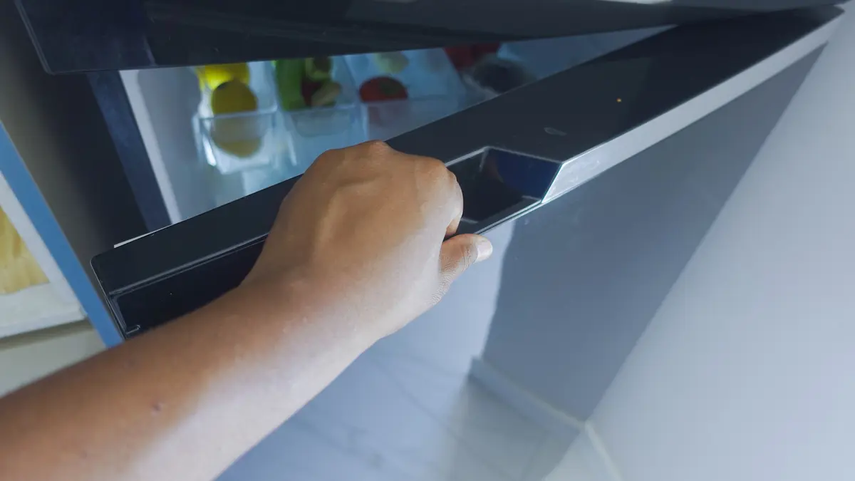 Hand opening a refrigerator door to store cooked pasta safely, avoiding counter cooling mistakes that cause harmful bacterial growth.