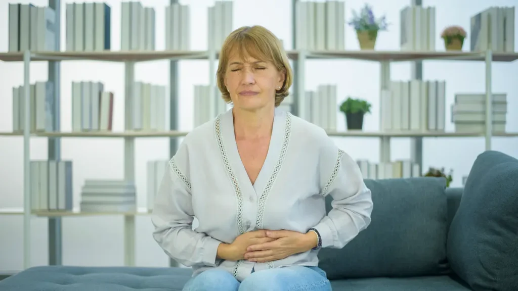Senior woman sitting on couch holding her stomach in pain, experiencing bloating or digestive discomfort after eating.