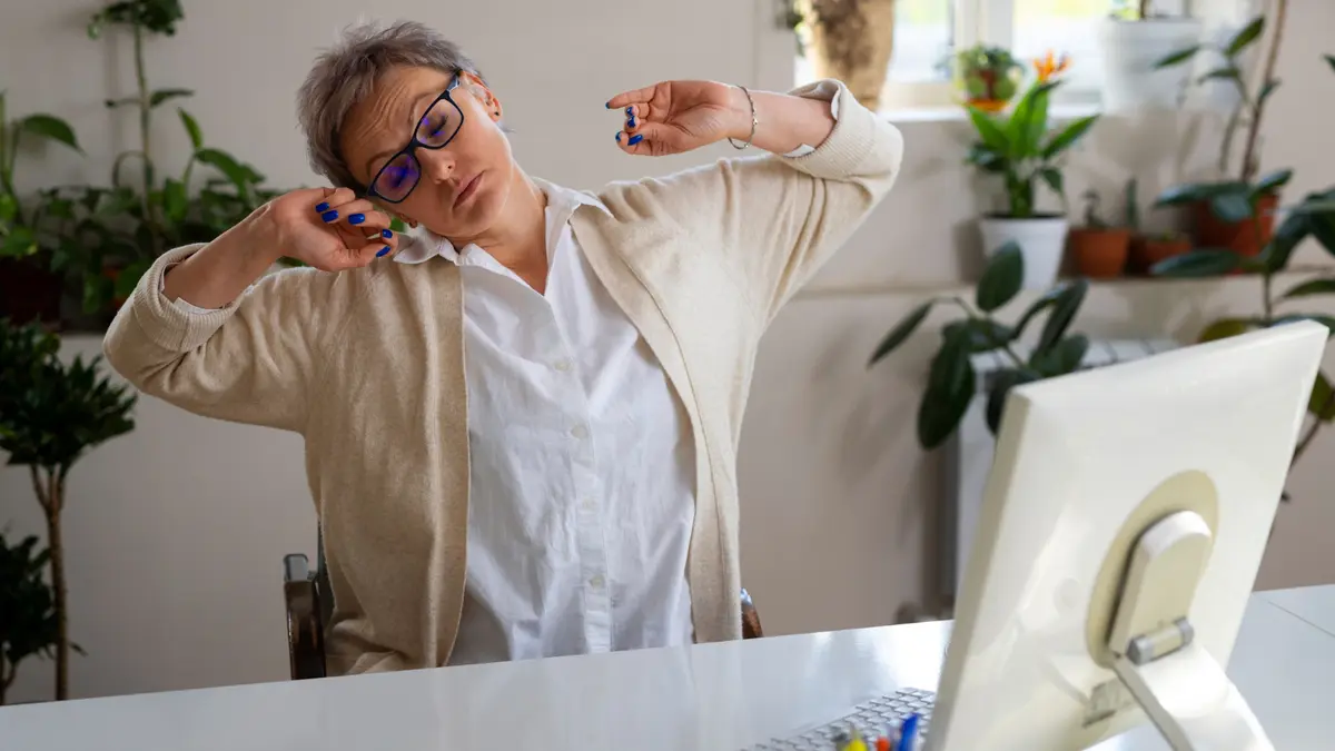 Silver-haired woman in her mid-50s stretching at a desk with heavy-lidded eyes, fighting the mid-afternoon energy crash that follows a low-fiber lunch.