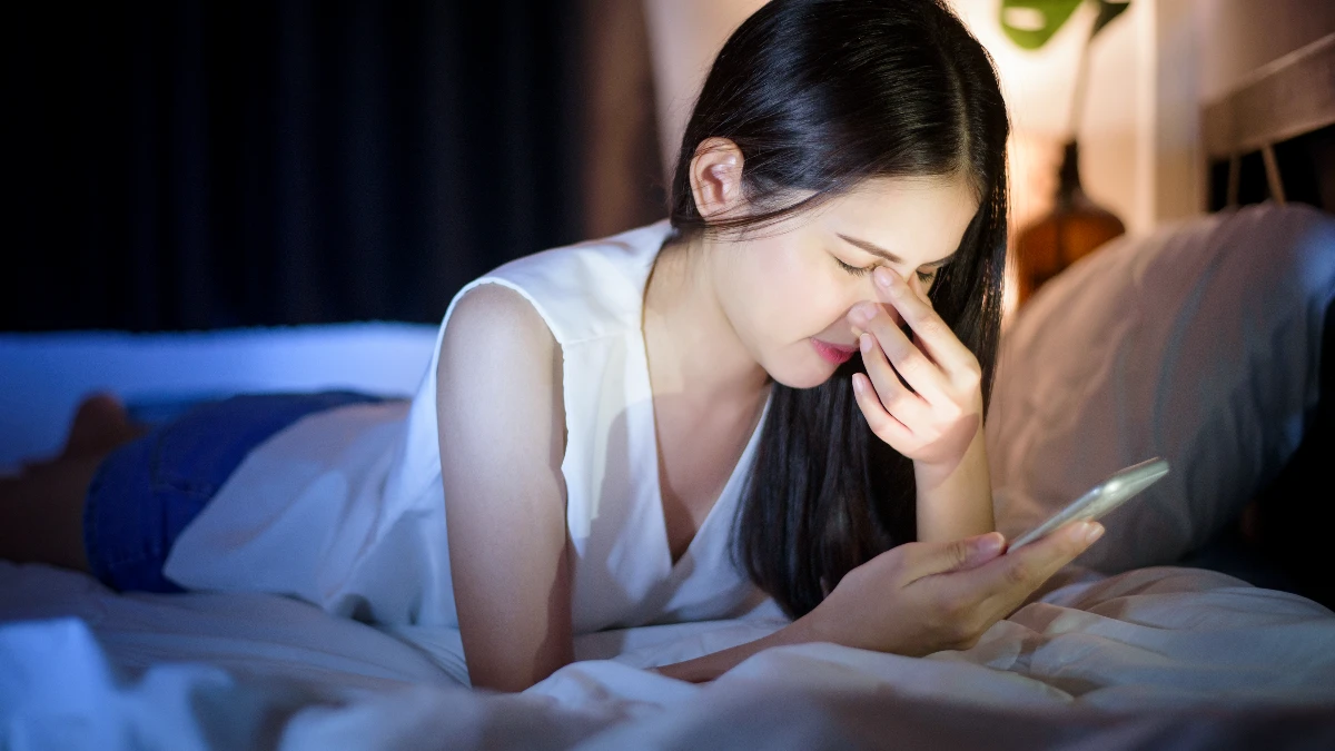 Exhausted woman lying in a dark bedroom rubs her strained eyes under the harsh blue glow of a smartphone, a habit that suppresses melatonin production and disrupts baseline cortisol management for the following morning.
