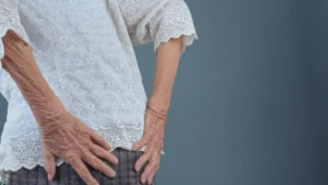 Close-up of an elderly woman in a white lace blouse holding her lower back in discomfort, illustrating the difficulty of standing up straight due to psoas-related stiffness.