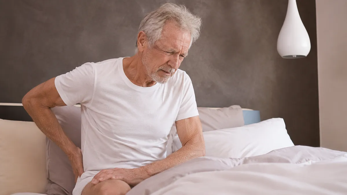 Older man in his 50s sitting on edge of bed gripping lower back with painful expression after morning soreness from exercise.