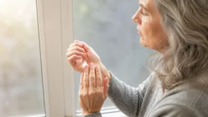 Older woman gently massaging stiff finger joints by a window in soft morning light, examining early signs of hand inflammation.