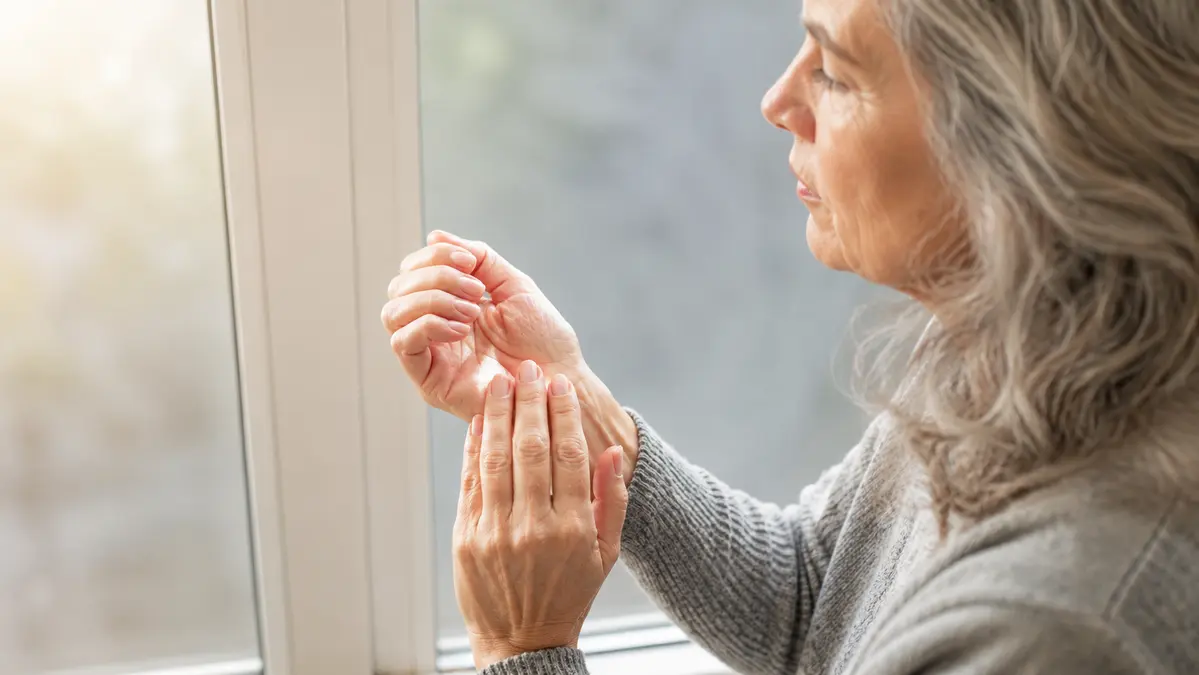 Older woman gently massaging stiff finger joints by a window in soft morning light, examining early signs of hand inflammation.