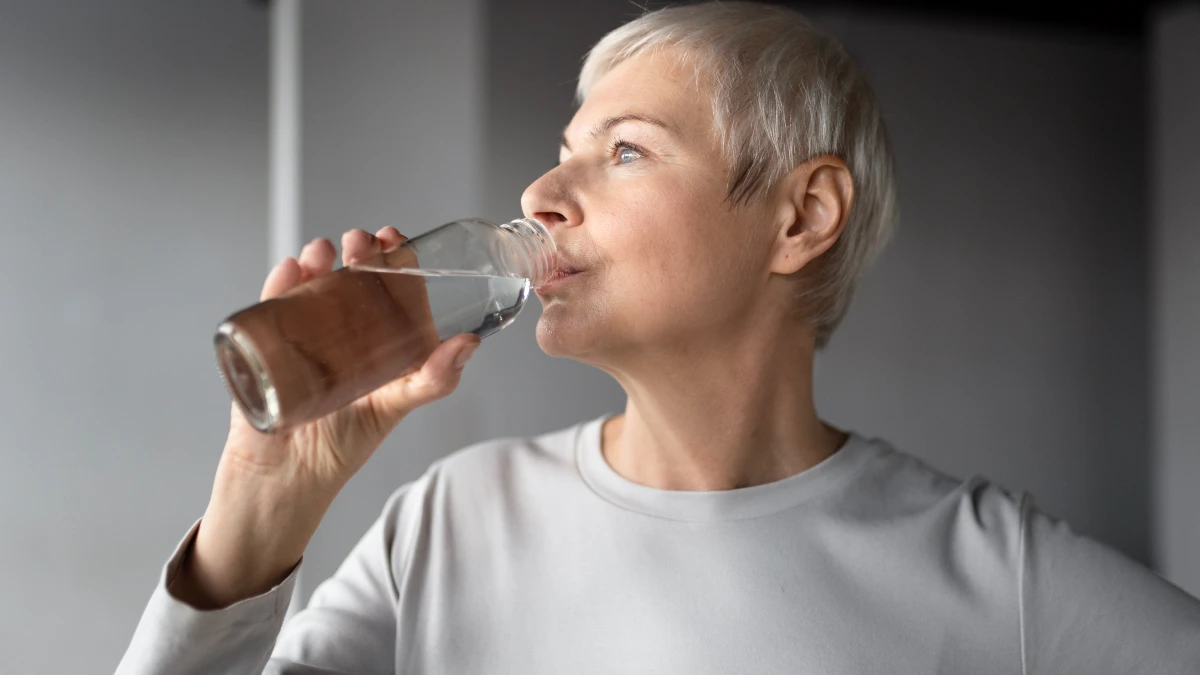 A close-up of an older woman with short silver hair wearing a light grey long-sleeved athletic shirt. She is looking off to the side while taking a sip of water from a clear glass bottle.