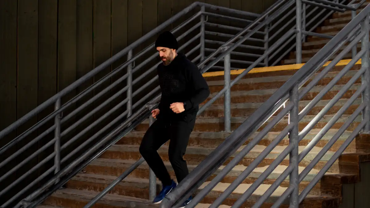 Man wearing a beanie and black athletic gear performing a step-down on outdoor wooden stairs, focusing on controlled movement to improve knee stability.