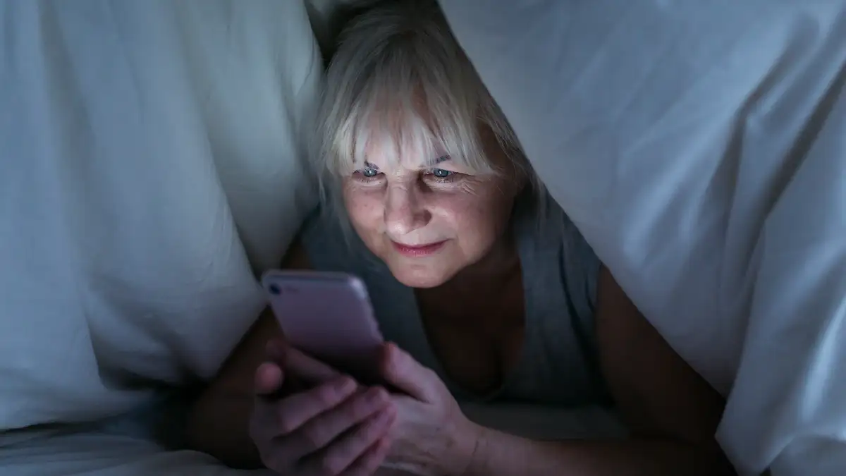 Mature woman lying under her covers in a dark bedroom stares at the harsh blue light of a smartphone, a casual late-night habit that suppresses sleep quality and disrupts her morning cortisol baseline.