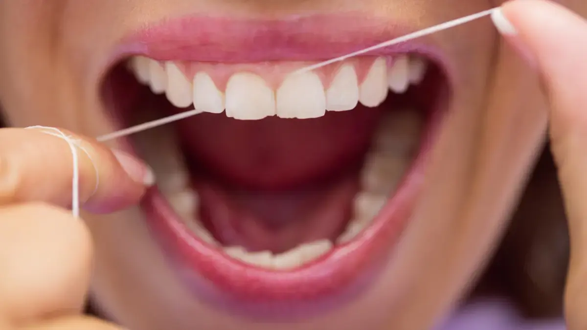 A close-up focuses on a person's open mouth with bright white teeth as they use a piece of dental floss, illustrating proper flossing technique to remove bacteria.