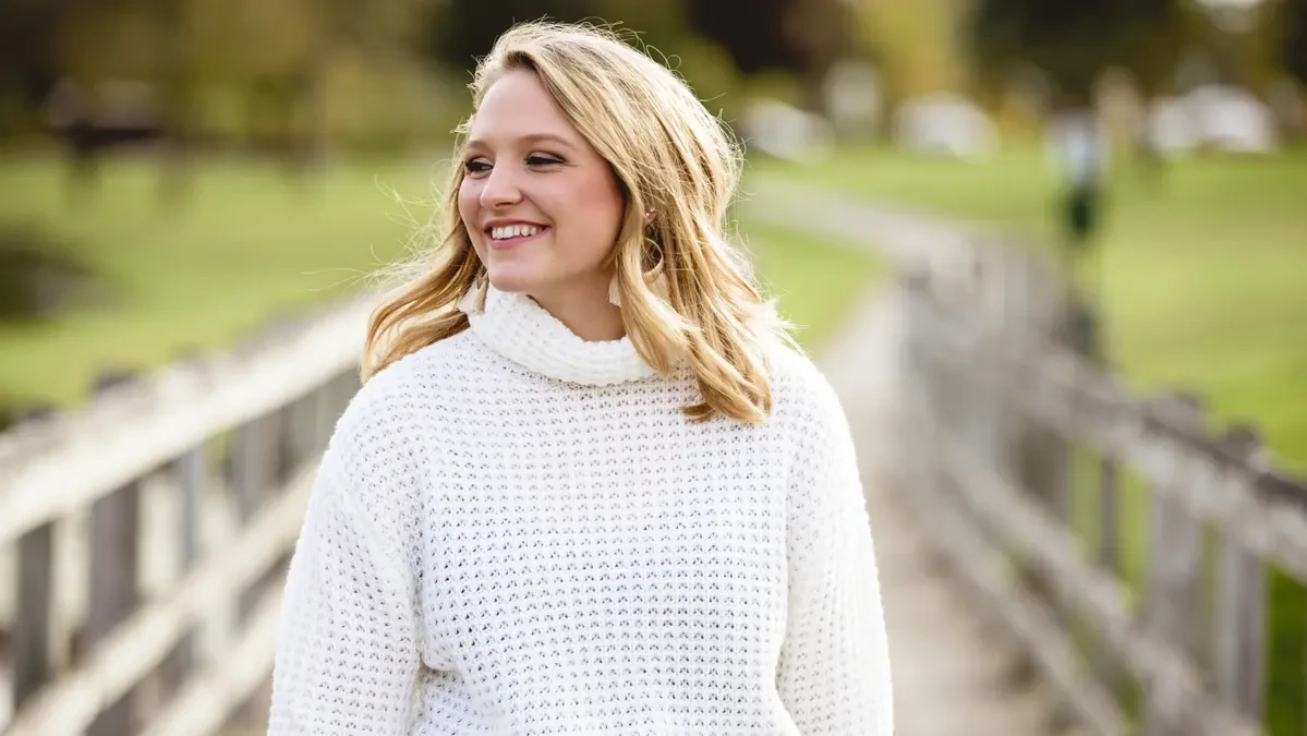 A smiling young woman with blonde hair wearing a white knit sweater stands outdoors on a wooden walkway, looking off to the side in soft morning light with a blurred green park in the background.