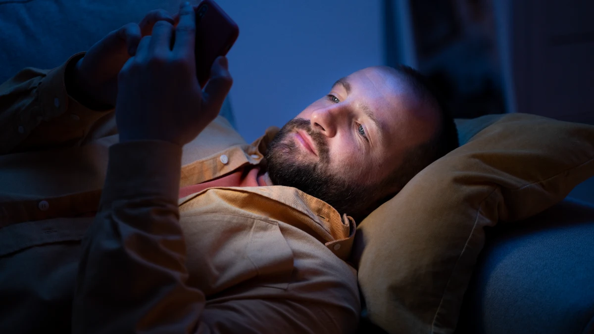 Man lying on a sofa in a dark room is illuminated by the harsh blue light of his smartphone, a casual evening habit that suppresses melatonin and disrupts the body's morning cortisol recovery.