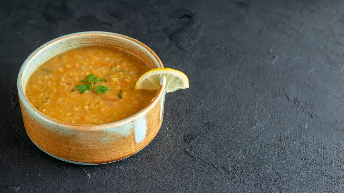 A rustic ceramic bowl filled with thick red lentil soup, topped with a fresh parsley garnish and a lemon slice on the rim, set against a dark textured background.