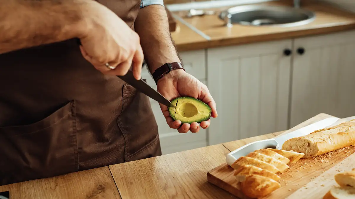 Man in a brown apron slicing a fresh avocado on a wooden counter to maintain proper electrolyte balance and support healthy muscle nerve-signaling.