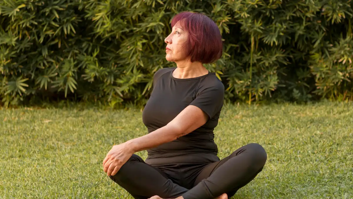 Mature woman sitting on a sunlit lawn performs a gentle seated spinal twist, using light outdoor movement to metabolize circulating stress hormones and support proactive cortisol management.