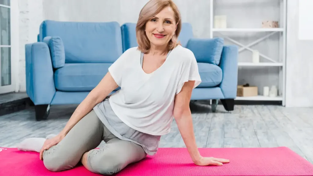 A smiling mature woman in her 60s sitting comfortably on a bright pink yoga mat in a modern living room, dressed in activewear and ready for a home stretching or foam rolling session.