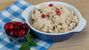 A bowl of creamy oatmeal topped with fresh red cranberries and a side of extra berries on a blue checkered napkin.