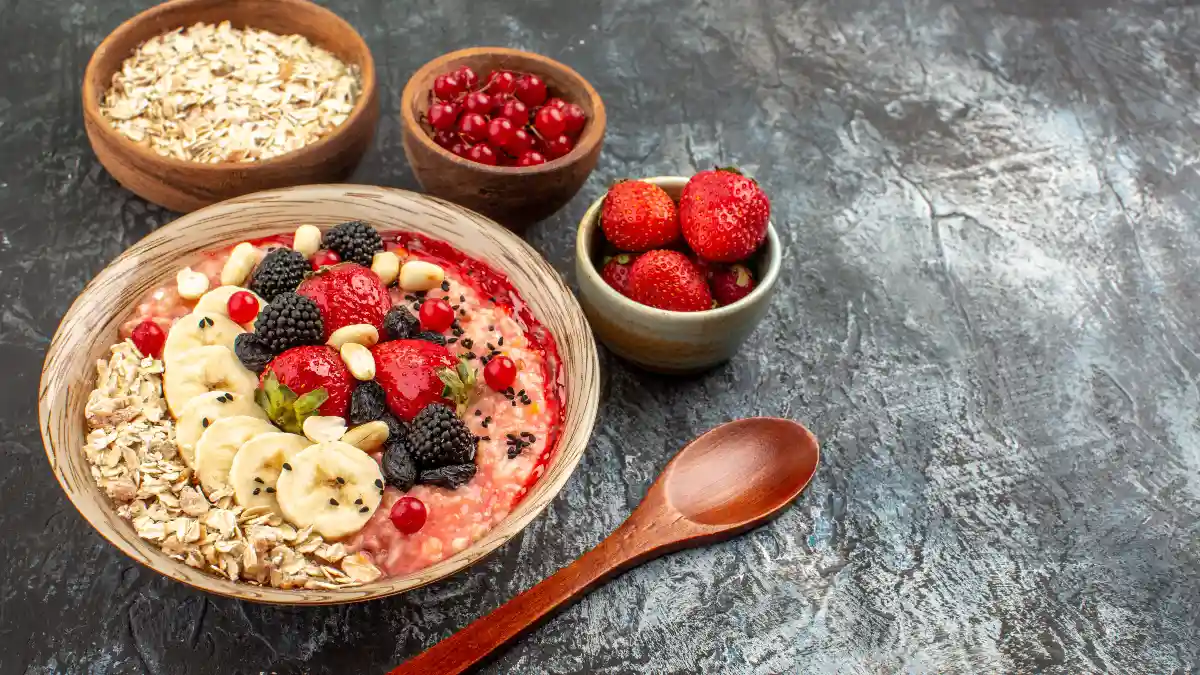 A vibrant bowl of oatmeal topped with sliced bananas, fresh strawberries, blackberries, and seeds, served with a wooden spoon on a dark textured tabletop.