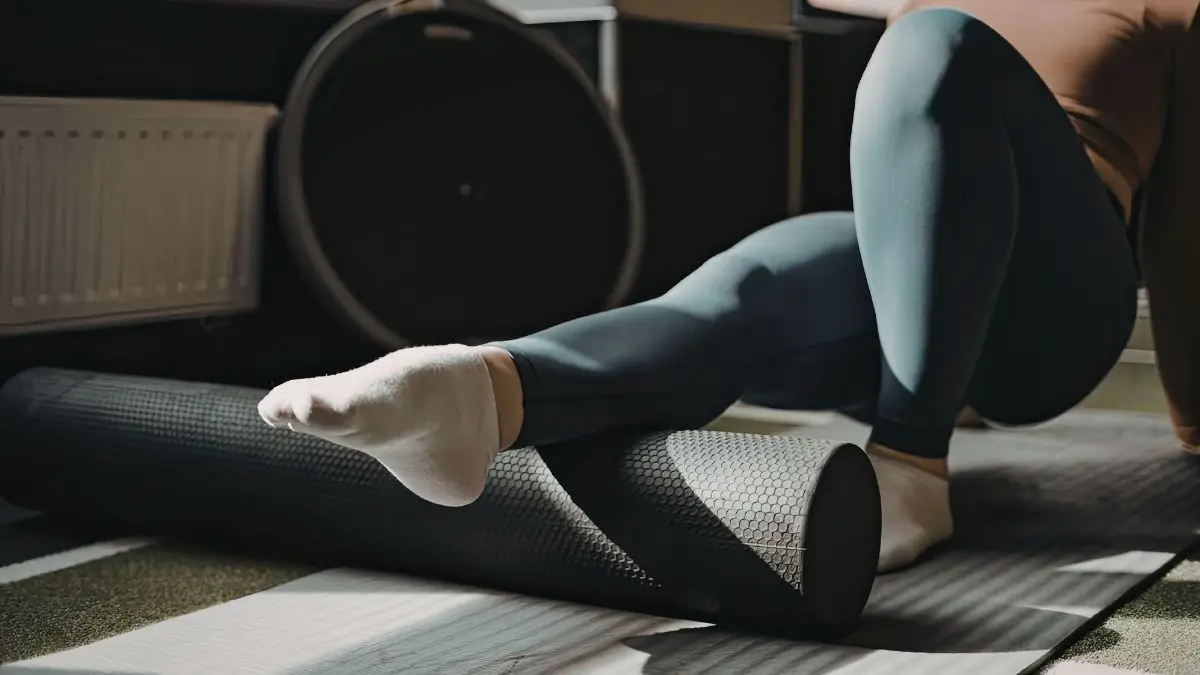 Close-up of a person’s legs in dark leggings and white socks using a black textured foam roller on a grey yoga mat to massage the calf muscle and release tension.