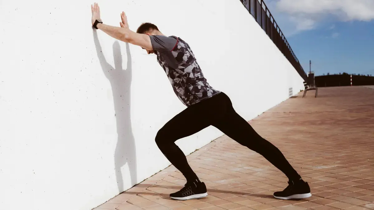 Adult male performing a wall-supported lunge with bent knees to target the soleus muscle and increase ankle range of motion as part of a mobility routine.