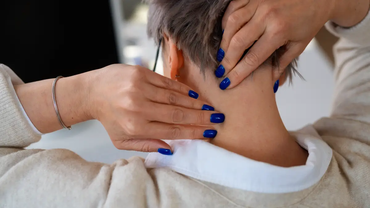 A mature woman massages the base of her neck to soothe muscle tension associated with hyperkyphosis after 50.
