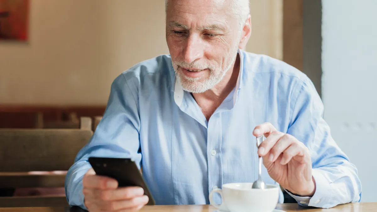 Mature man checking his smartphone while drinking coffee to set a daily reminder that supports his personal caffeine clearance and sleep quality.