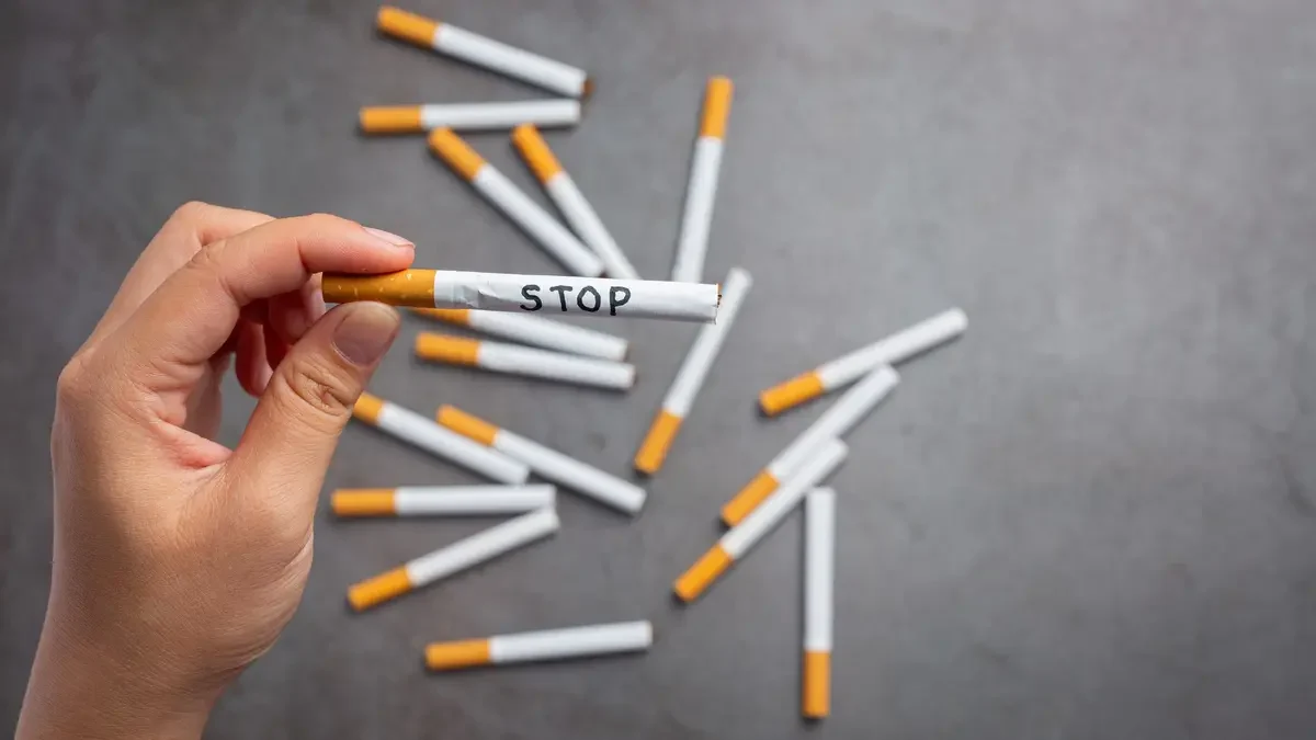 A top-down view of a hand holding a single cigarette with the word STOP handwritten in black ink on the white paper. In the background, several other cigarettes are scattered haphazardly across a dark, textured gray surface, out of focus