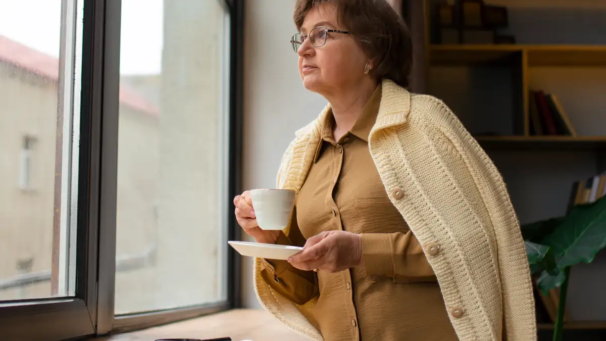 Thoughtful mature woman stands by a bright window holding a warm cup, strategically delaying her morning caffeine intake to prevent an artificial surge during her peak cortisol window.