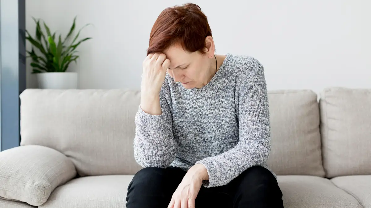 Distressed mature woman in a grey sweater sits on a light sofa resting a hand against her forehead, physically enduring the unexplained morning dread triggered by a haywire biological event after fifty.