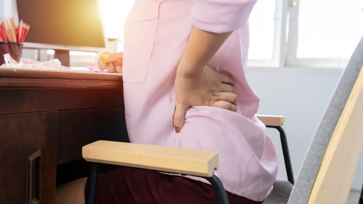 Mature woman slouched in a home office chair with hips flexed to illustrate how sitting too long causes psoas muscle shortening and chronic lower back stiffness.