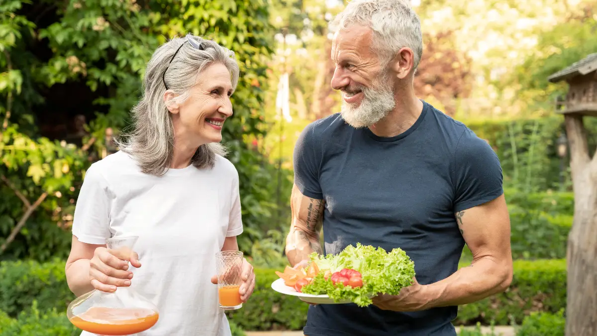 Joyful mature couple laughing together in a sunlit garden while sharing a plate of fresh greens and tomatoes to support a healthy, longevity-focused lifestyle