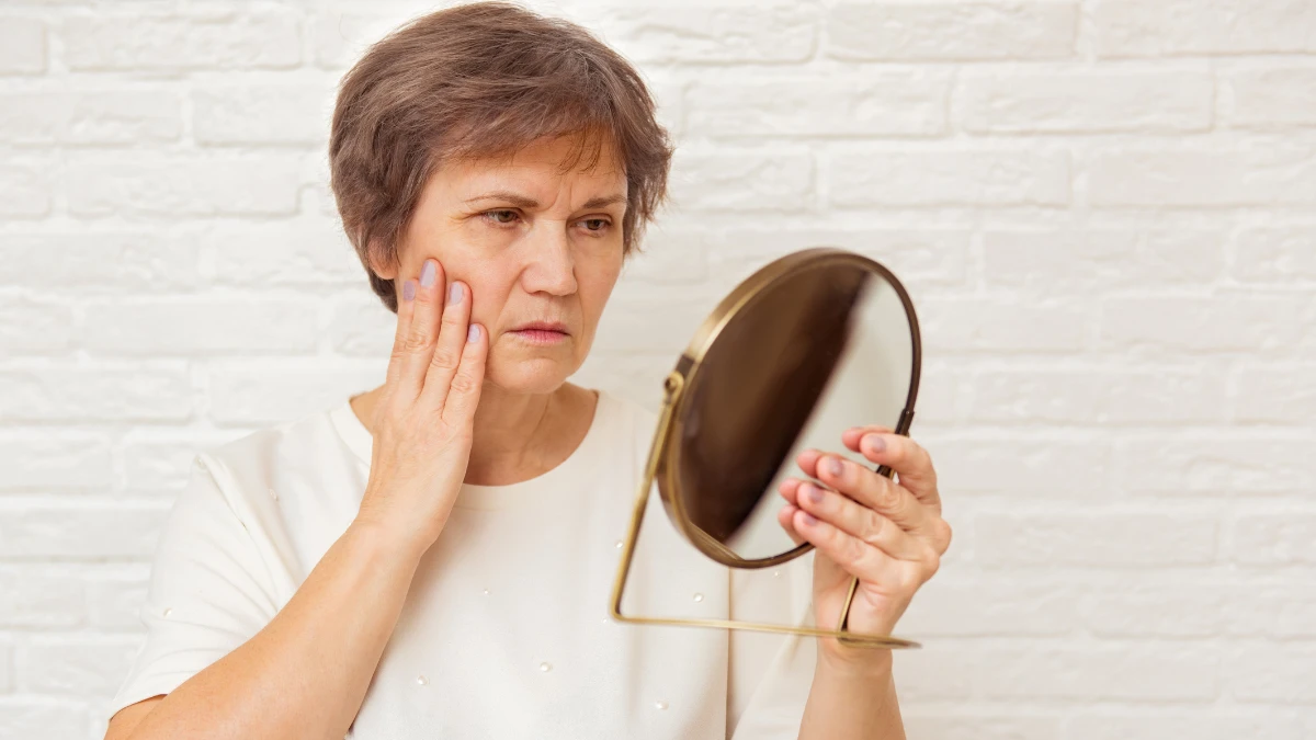 Mature woman inspecting her gum line in a sunlit mirror to detect early signs of periodontitis after 50 and prevent permanent bone loss.