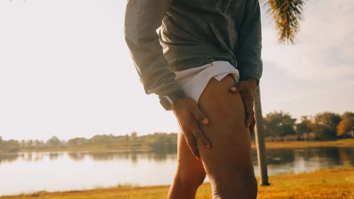 Close-up of a mature male runner pausing outdoors to hold his upper thigh in pain, illustrating the difference between localized psoas tension and radiating sciatic nerve symptoms.