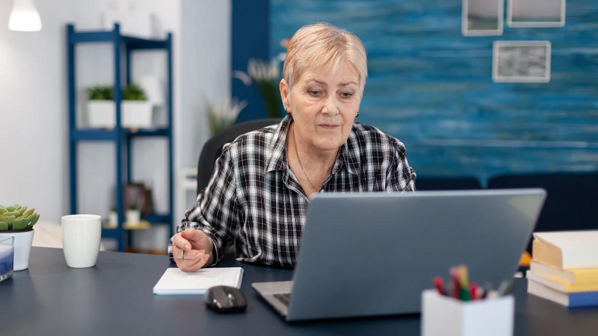 Focused mature woman in a plaid shirt sits at her home office desk looking at a laptop and holding a pen over a notebook, preparing a list of questions about hormone panels and proactive cortisol management to discuss with her physician.