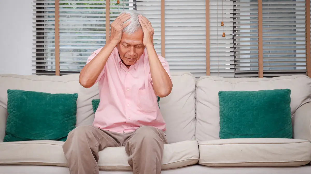 Senior Asian man sitting on a sofa holding his head in frustration, representing the mental fatigue and brain fog caused by chronic stress.