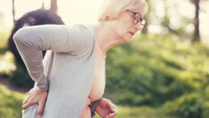 A woman over 50 in a gray cardigan holding her lower back with a look of discomfort, illustrating the physical hesitation and unsteadiness common with aging.
