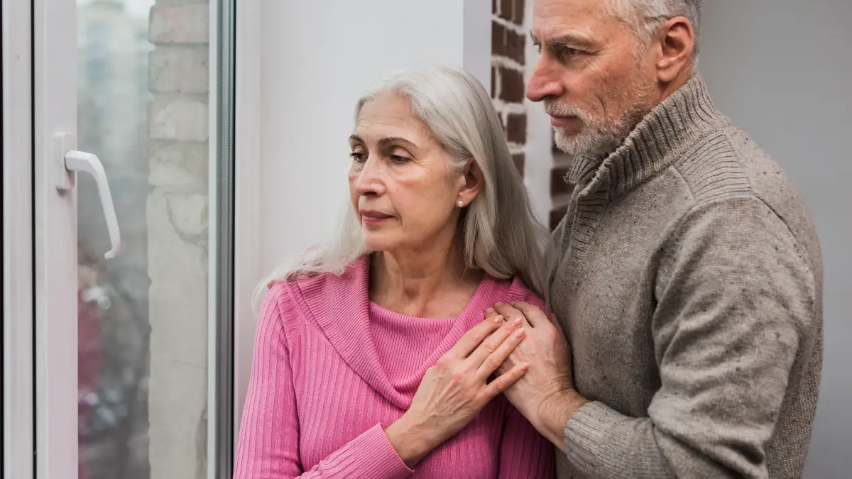 Mature couple looking out a window at dusk to avoid navigating low-light environments that challenge visual contrast sensitivity.