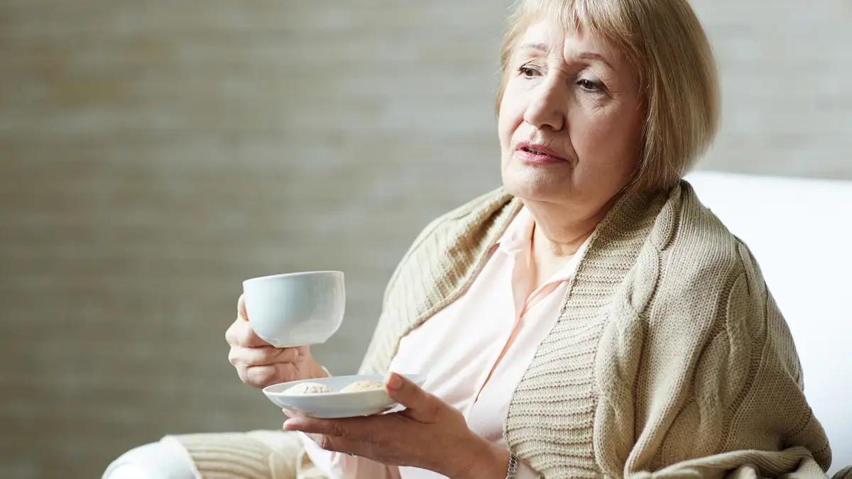 Senior woman wrapped in a warm shawl holding a cup of tea to illustrate the slow recovery and fatigue caused by a weakened immune system in older adults.