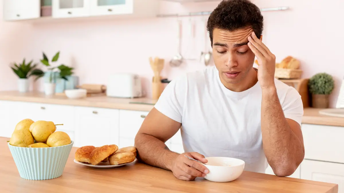 Young man rubbing his forehead in fatigue while sitting at a kitchen counter with food, illustrating the post-meal sluggishness that worsens as the gut microbiome ages.