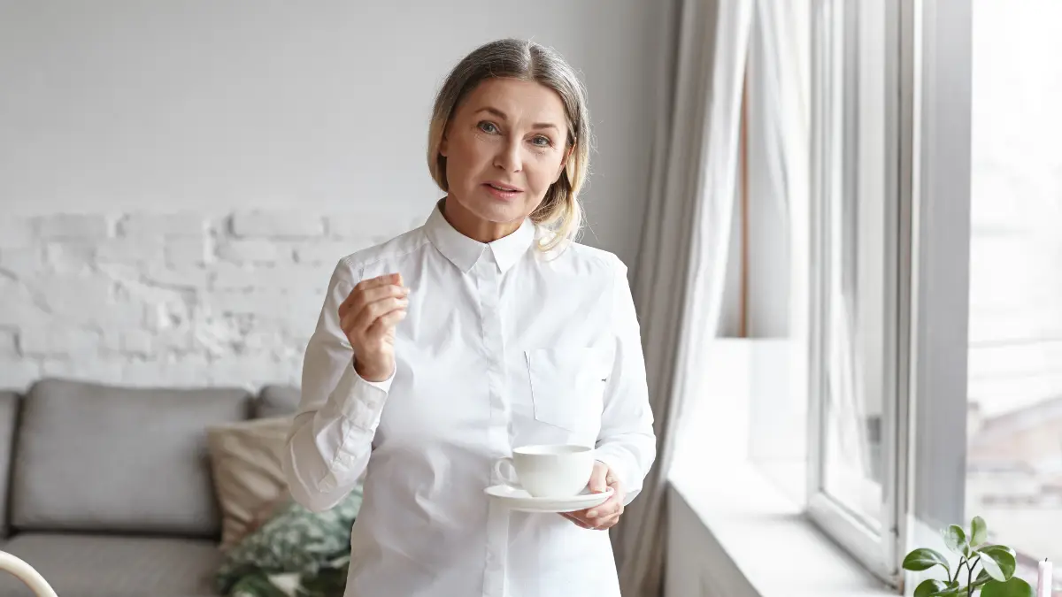 Mature woman in a white button-down shirt holding a ceramic teacup and saucer to boost metabolism naturally during her morning routine.