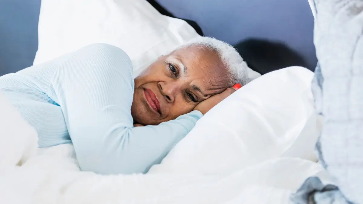 Mature woman lying awake on white linens with a contemplative expression to improve cortisol management and encourage deeper sleep cycles.