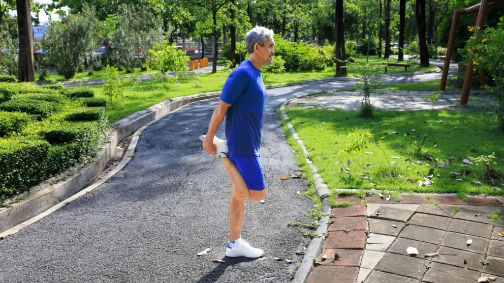 Active senior man with silver hair wearing a blue workout kit stretches his quad on a paved park path to maintain the muscle mass and fitness needed for good balance.