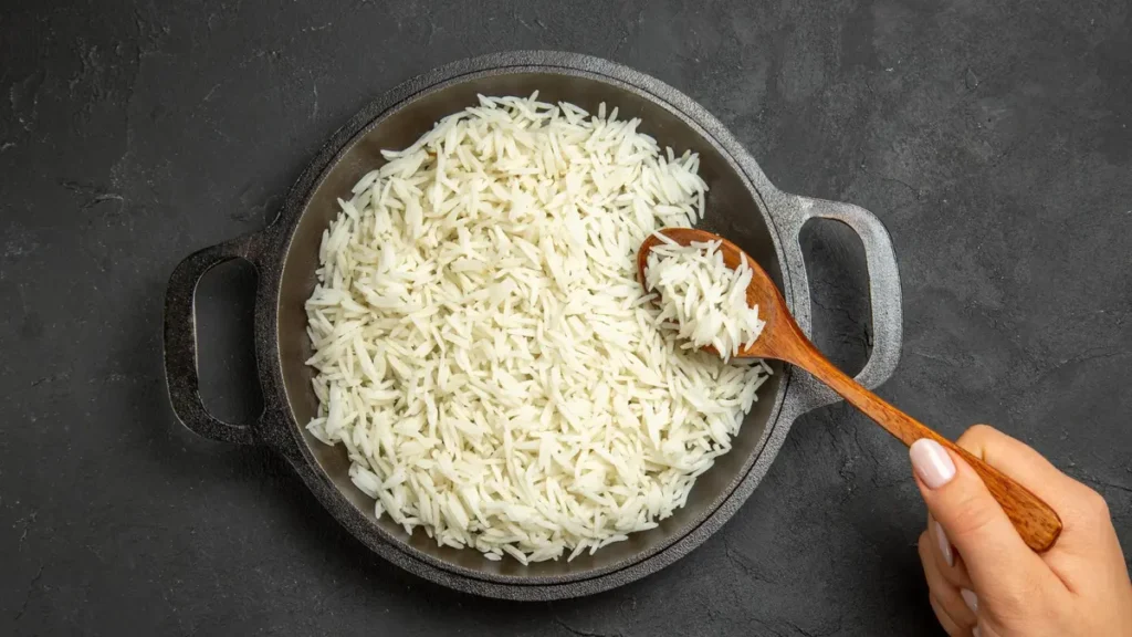 A cook uses a wooden spoon to spread cooked long-grain basmati rice in a flat pan to cool down before refrigerating it.