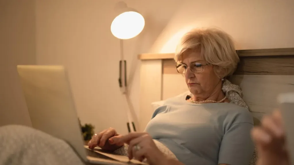 Senior woman using laptop in bed at night with bright bedside lamp — screen light and room lighting disrupting sleep after 50.
